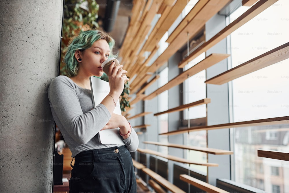 Alternative girl in casual clothes and with green hair standing indoors at daytime with cup of drink in hands.