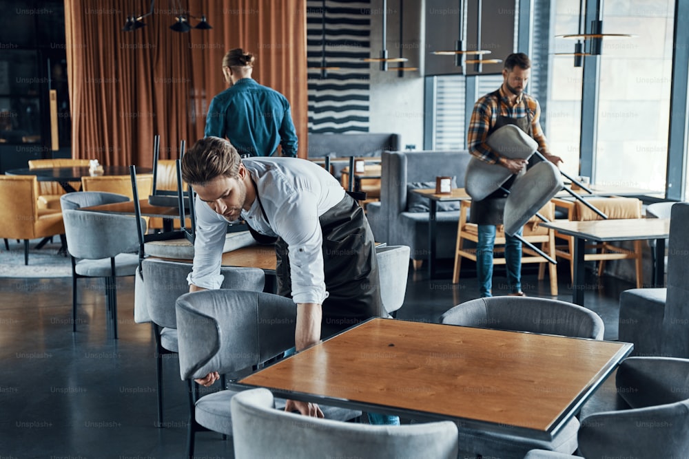 Hübsche junge Männer in Schürzen arrangieren Möbel, während sie das Restaurant bis zur Eröffnung vorbereiten