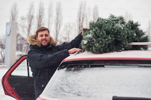 Young man standing near car with green christmas tree on top.
