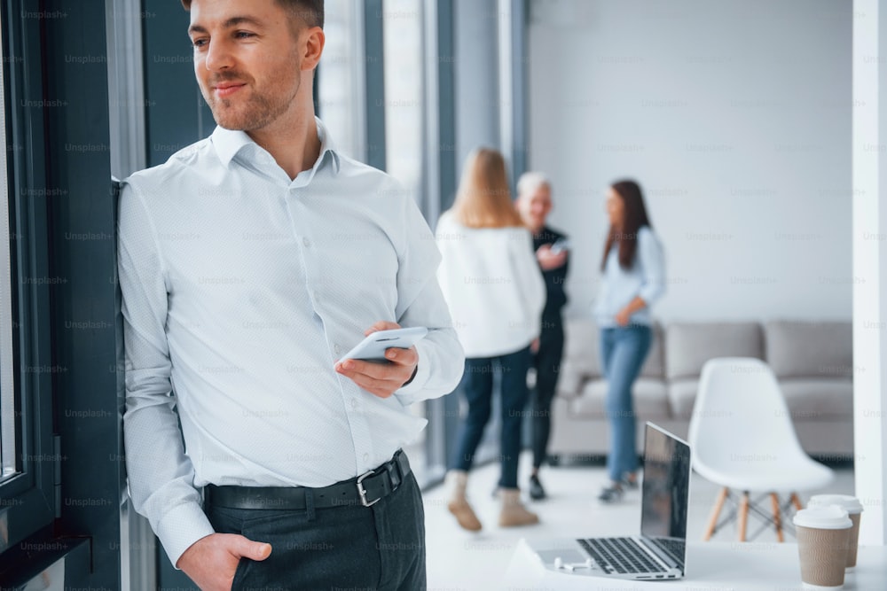Homem em roupas formais em pé com o telefone na mão na frente de uma jovem equipe de sucesso que trabalha e se comunica em conjunto dentro de casa no escritório.