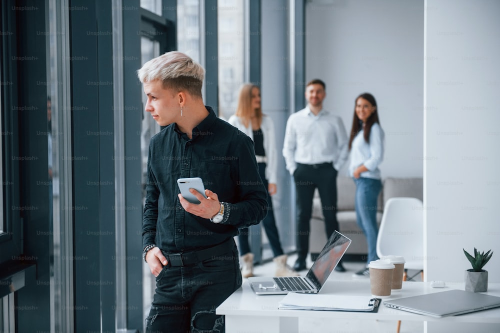 Young hipster with phone standing in front of group of young successful team that working and communicating together indoors in office.