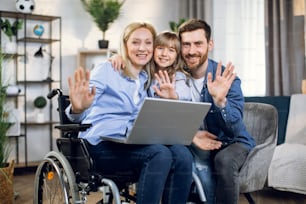 Behinderte Frau mit Laptop auf den Knien, die ihre geliebte Tochter und ihren geliebten Ehemann umarmt. Glückliche junge Familie, die die gemeinsame Zeit zu Hause genießt. Menschen mit Behinderungen.