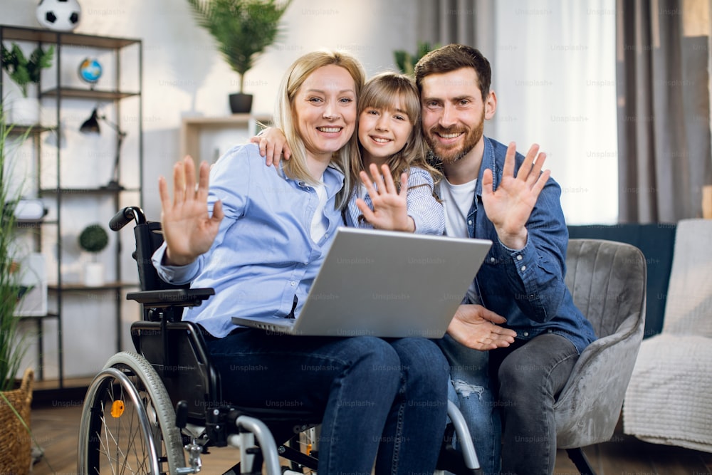 Mujer discapacitada con computadora portátil de rodillas abrazando a su amada hija y esposo. Familia joven y feliz disfrutando del tiempo juntos en casa. Personas con discapacidad.