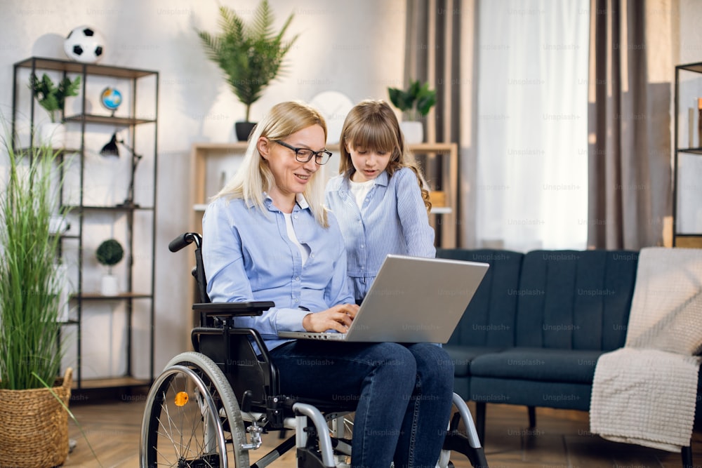 Pretty daughter embracing handicapped mother that sitting in wheelchair with modern laptop on knees. Concept of people, technology and disability.