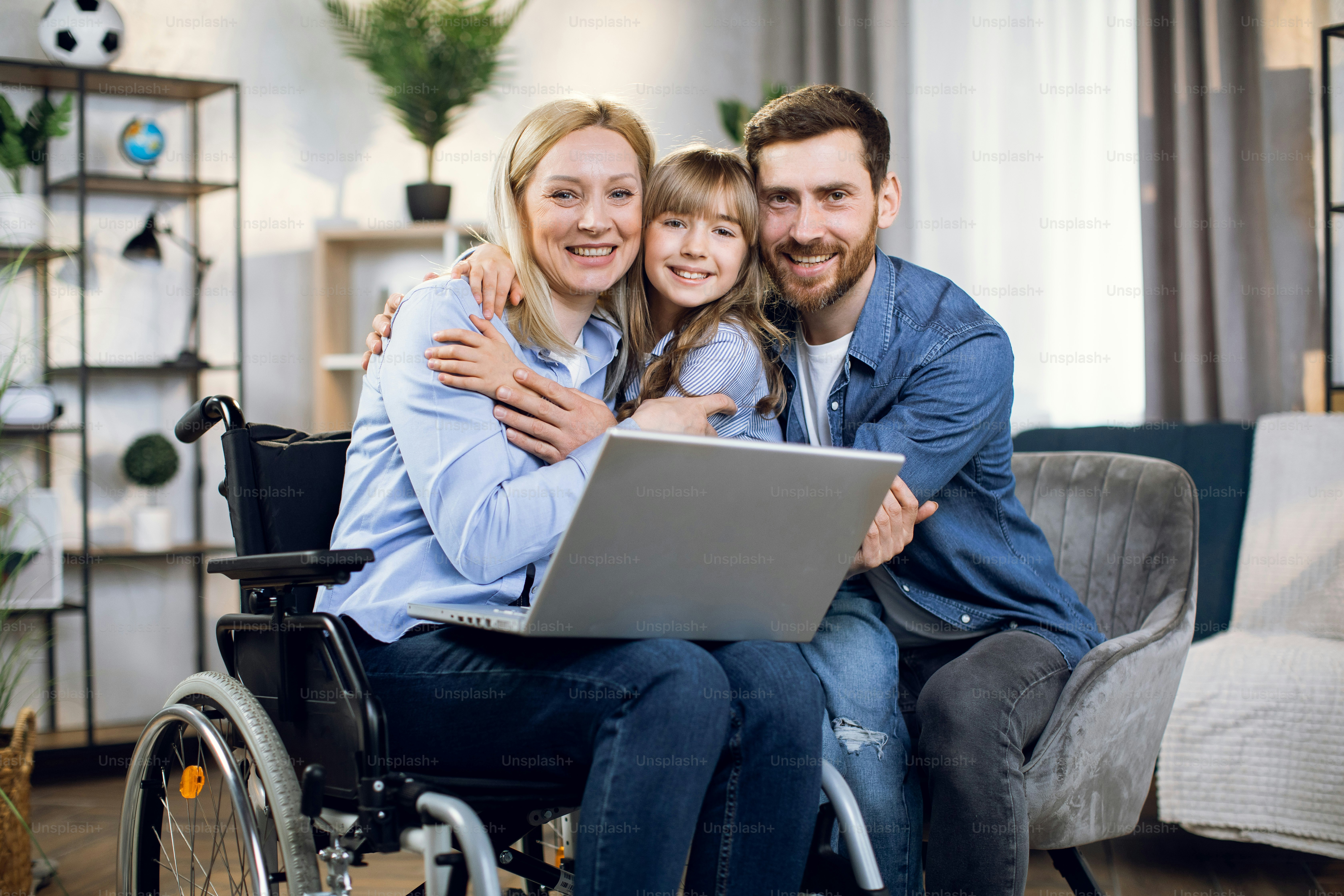 Femme handicapée avec un ordinateur portable sur les genoux embrassant sa fille et son mari bien-aimés. Jeune famille heureuse profitant du temps ensemble à la maison. Personnes en situation de handicap.