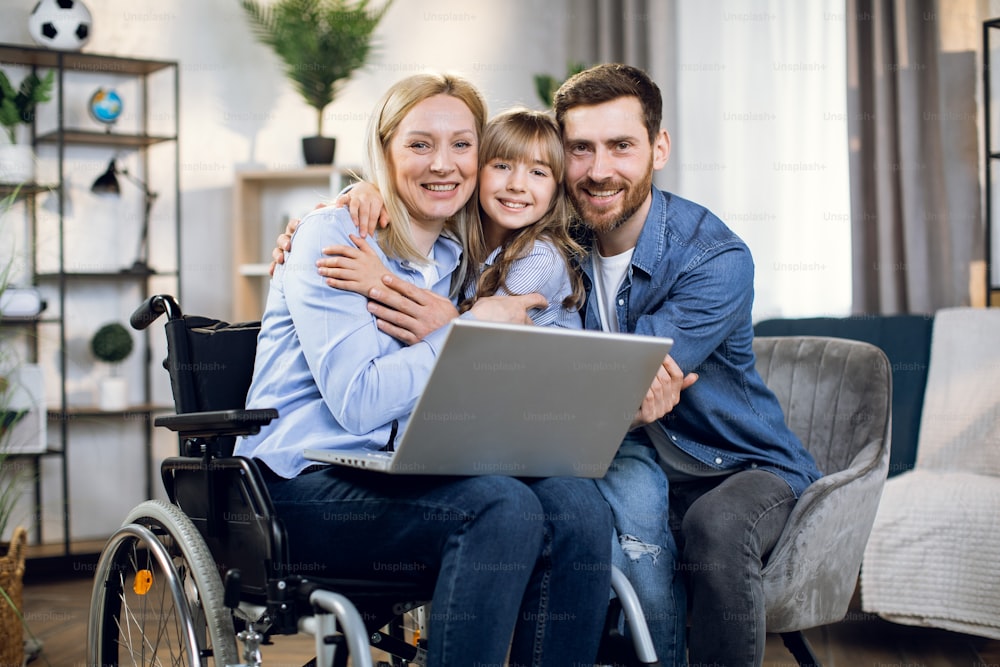 Handicapped woman with laptop on knees embracing her beloved daughter and husband. Happy young family enjoying time together at home. People with disabilities.
