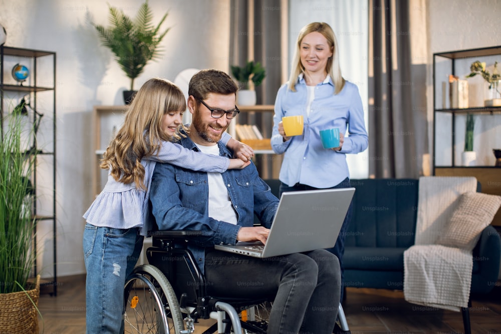 Smiling woman holding two cups of coffee while standing near disabled husband that hugging cute daughter. Handicapped man working remotely on laptop to support family.