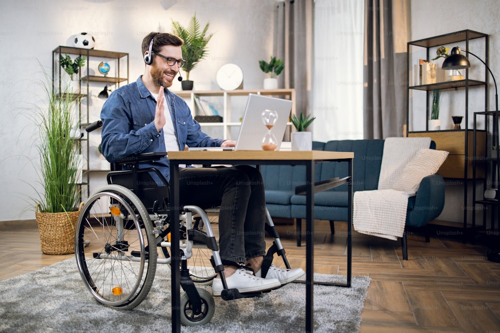 Hombre barbudo con anteojos y auriculares hablando y gesticulando durante una videoconferencia en una computadora portátil moderna. Freelancer con discapacidad que tiene la oportunidad de trabajar desde casa.