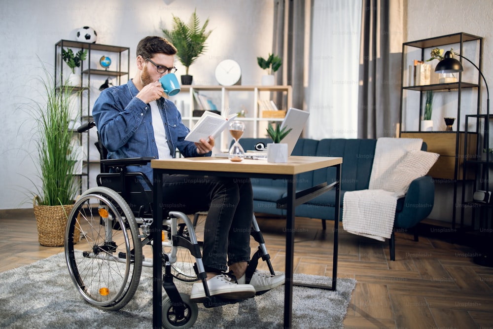 Handicapped young man drinking coffee and reading book while while sitting at table with modern laptop. Male freelancer in wheelchair relaxing during remote work at home.