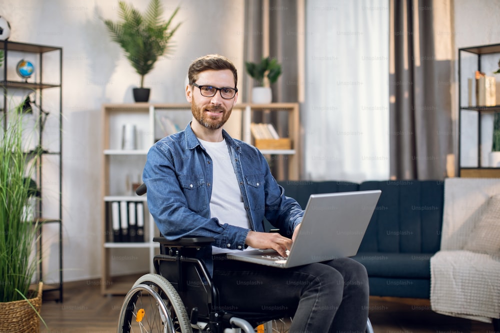 Young male freelancer sitting in wheelchair, using wireless laptop and looking at camera. Remote work for disabled people. Technology concept.