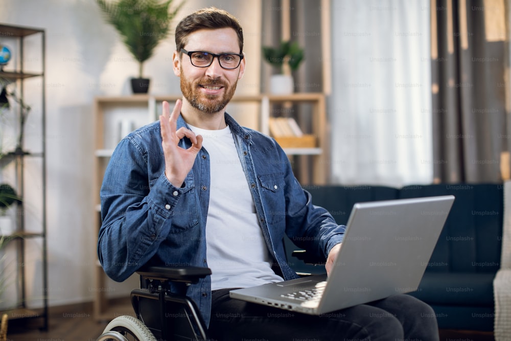 Handsome bearded guy with disabilities smiling and gesturing on camera while sitting in wheelchair with modern laptop. Positive mood of handicapped freelancer.