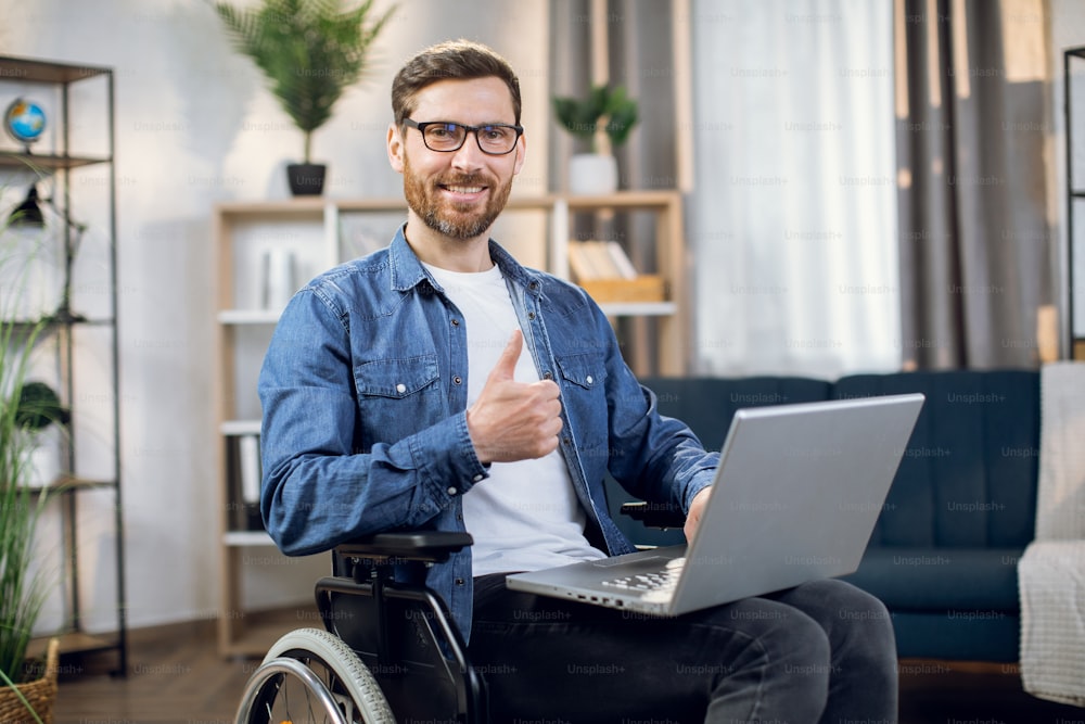 Hombre barbudo sonriente mostrando el pulgar hacia arriba mientras está sentado en silla de ruedas y sostiene una computadora portátil inalámbrica sobre las rodillas. Feliz autónomo discapacitado que trabaja desde casa.