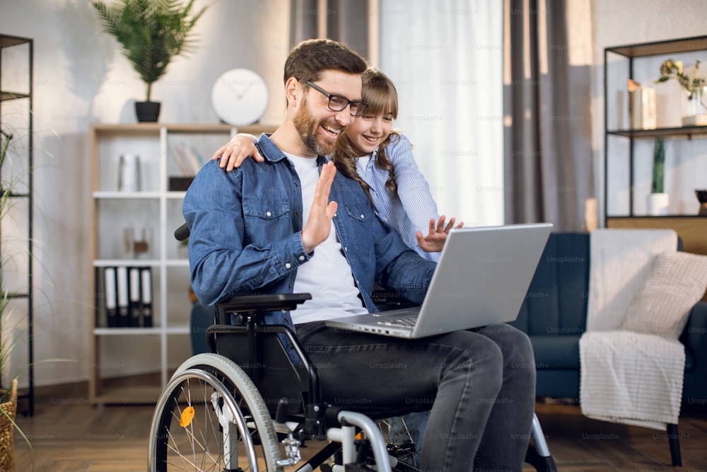 Uomo disabile barbuto e sua figlia carina che sorridono e salutano durante la videochiamata sul laptop moderno. Maschio seduto su sedia a rotelle, femmina in piedi vicino a lui e abbracciato.