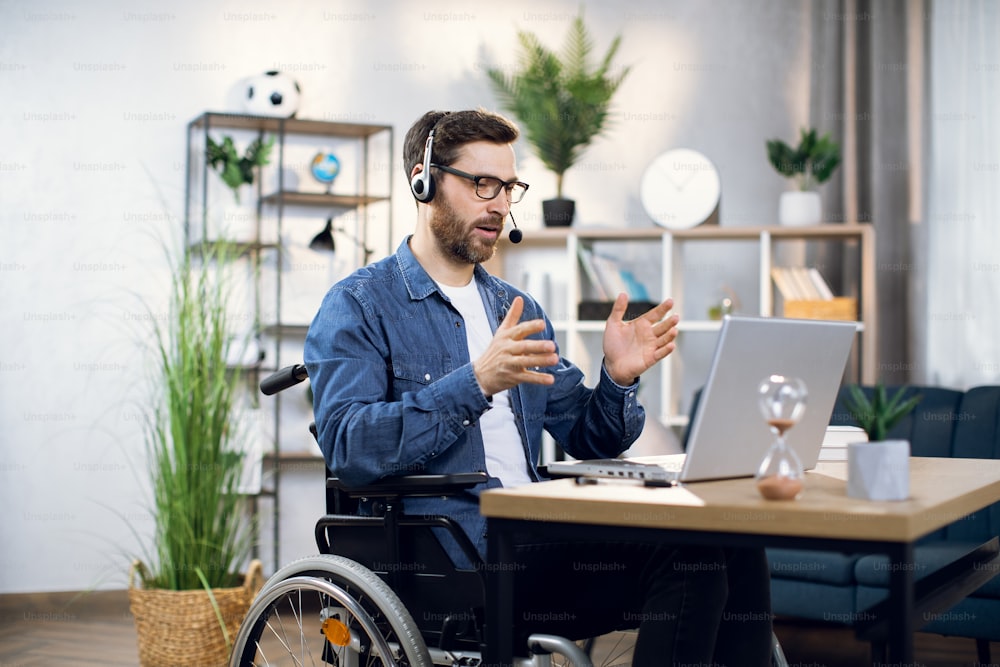 Bearded man in eyeglasses and headset talking and gesturing during video conference on modern laptop. Freelancer with disabilities having opportunity to work from home.