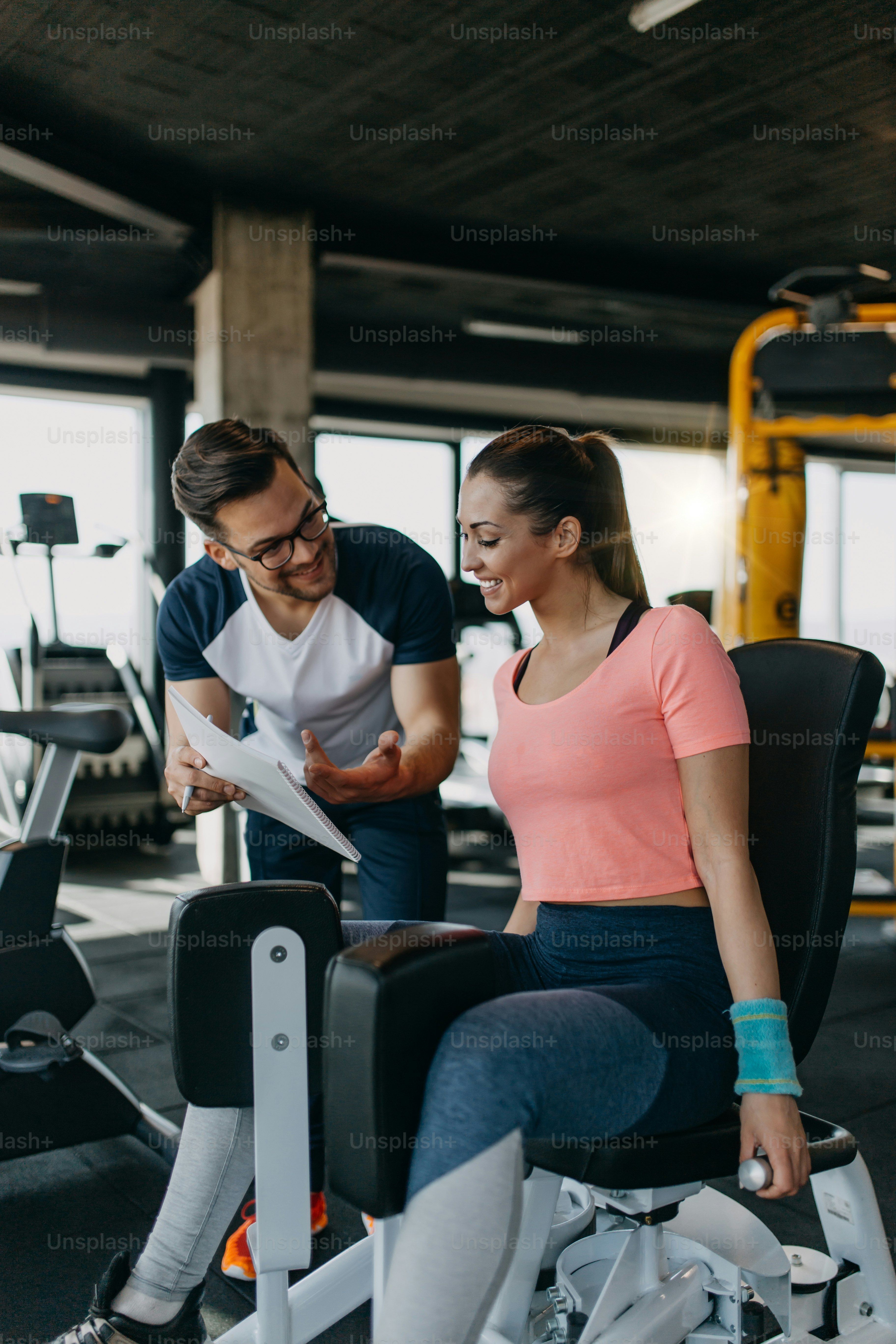 Wanita muda yang bugar dan menarik berolahraga di gym modern bersama dengan instruktur atau pelatih kebugaran pribadinya.
