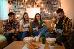 A group of friends on a house party playing guitar and drinking beer while having fun and eating nachos and popcorn