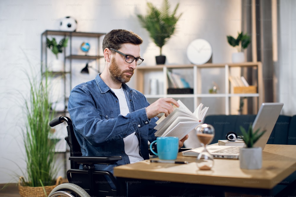 Bärtiger junger Mann im Rollstuhl liest Buch während der Kaffeepause zu Hause. Männlicher Freiberufler mit Behinderungen sitzt am Arbeitsplatz mit drahtlosem Laptop.