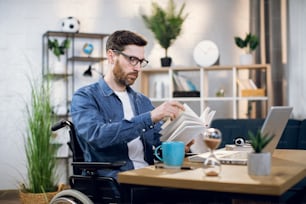 Jeune homme barbu en fauteuil roulant lisant un livre pendant la pause-café à la maison. Pigiste handicapé assis sur son lieu de travail avec un ordinateur portable sans fil.