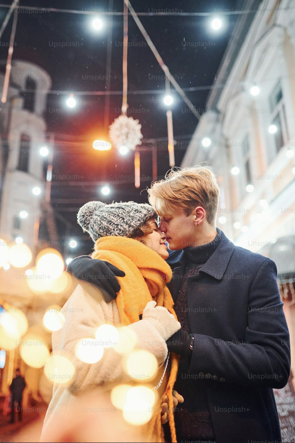 Junges Paar in warmen Kleidern küsst sich auf weihnachtlich geschmückter Straße.