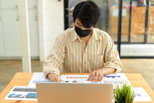 Porträt eines Geschäftsmannes mit Gesichtsmaske, der mit Papierkram und Laptop im Büroraum arbeitet