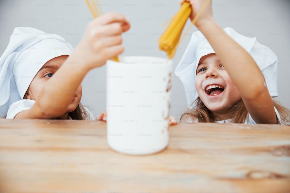 Two cute little girls in white chef hats holding spaghetti and puts it to big cup.