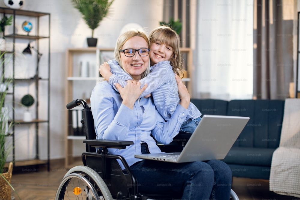Cheerful woman and pretty child posing in embrace at living room. Disabled mother with laptop on knees spending time at home with her daughter.