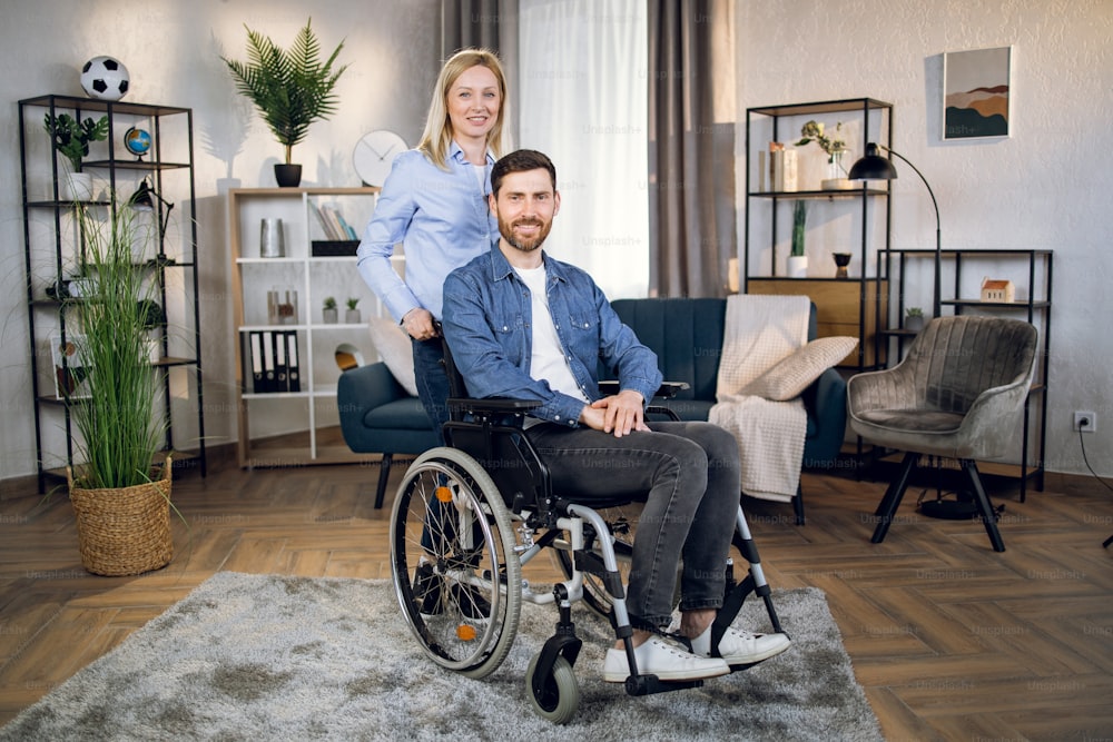 Attractive young woman smiling and looking at camera while standing near handsome man that sitting in wheelchair. Lovely wife helping and supporting disabled husband at home.