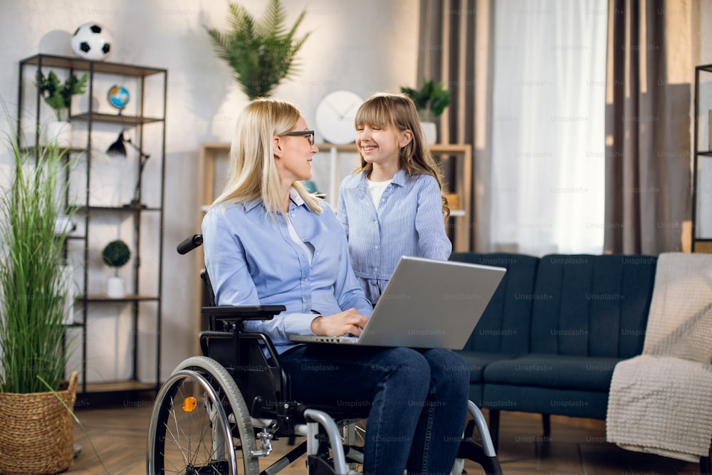 Attractive woman sitting in wheelchair with modern laptop and talking to her cute daughter. Casual day of disabled people. Technology concept.