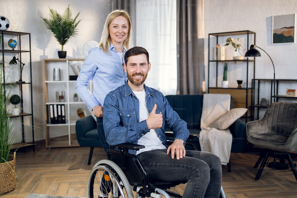 Hombre discapacitado sonriente sentado en silla de ruedas y mostrando el pulgar hacia arriba mientras su encantadora esposa con cabello rubio está de pie detrás. Personas con discapacidad y concepto de apoyo.