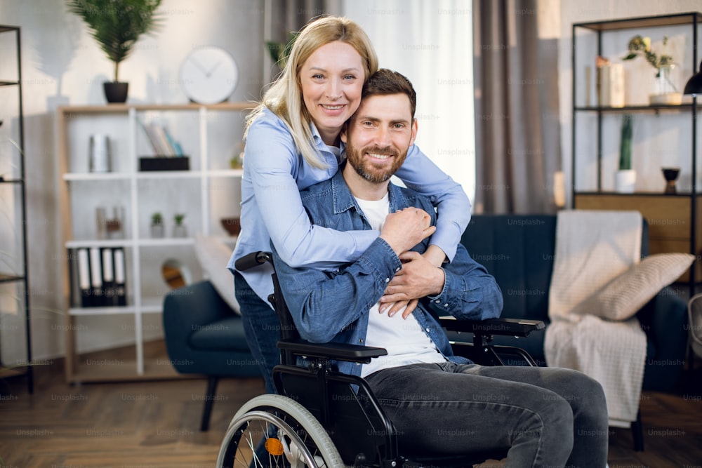 Retrato de una pareja casada feliz sonriendo y mirando a la cámara en la sala de estar. Bonita esposa abrazando a su marido discapacitado. Estado de ánimo positivo.
