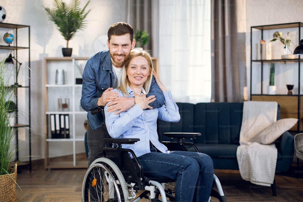 Beautiful woman with blond hair sitting in wheelchair while handsome husband standing behind and hugging her. Man supporting wife during rehabilitation. Relationship concept.