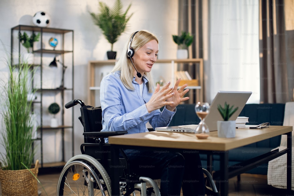 Pretty young woman in wheelchair using headset and laptop for video conference at home. Handicapped business lady leading working meeting online.