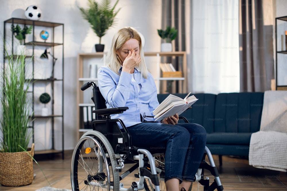 Attractive woman with blond hair sitting in wheelchair and rubbing eyes after long read of interesting book. Concept of disabled people, literature and leisure time.