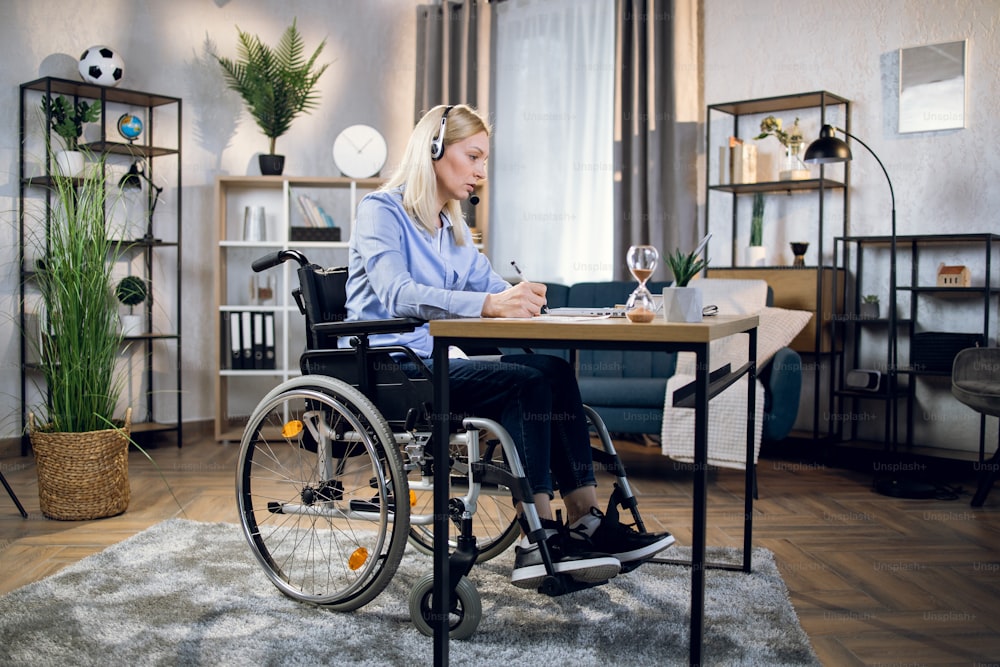 Side view of woman with disabilities taking notes during video conference at home. Female freelancer using headset and laptop for work. People with special needs.