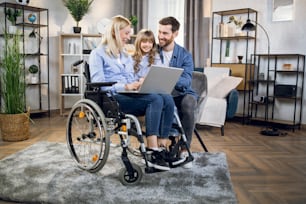 Pretty little kid with father and disabled mother using modern laptop during free time at home. Female blonde sitting in wheelchair. Casual day of young family.