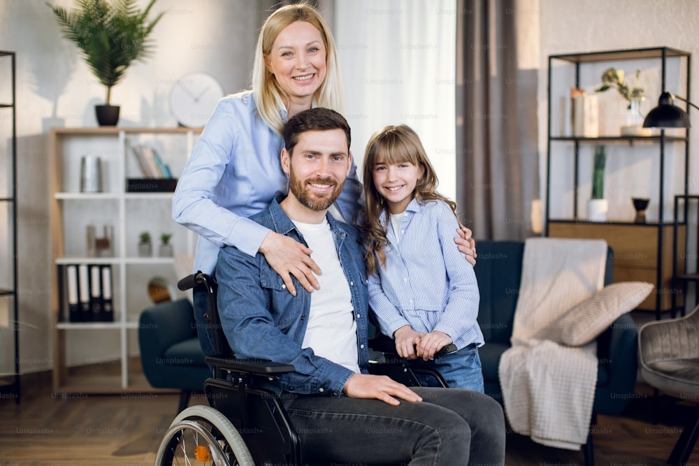 Disabled man sitting in wheelchair while his wife and daughter standing near and hugging him. Young family with one handicapped person.