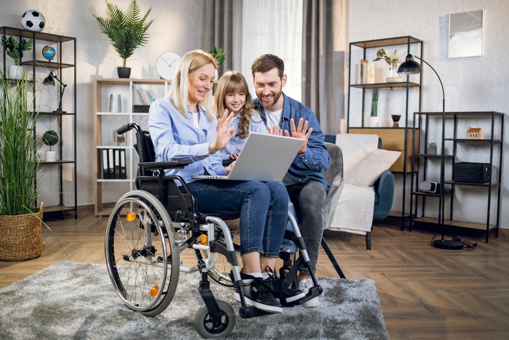 Handicapped woman, cute child and handsome man smiling and waving hands during video chat on modern laptop. Young parents with daughter enjoying online conversation while staying at home.