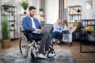 Bearded young man sitting in wheelchair with wireless laptop. Blur background of beautiful woman and cute child sitting on couch and using computer.