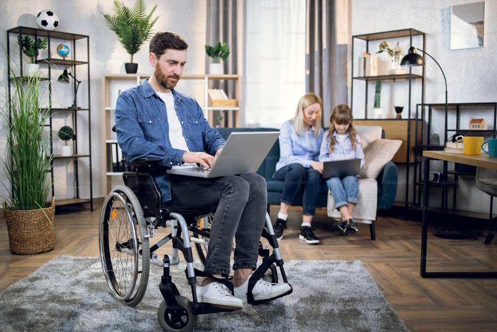 수염을 기른 청년이 무선 노트북으로 휠체어에 앉아 있다. 아름다운 여자와 귀여운 아이가 소파에 앉아 컴퓨터를 사용하는 배경을 흐리게 한다.