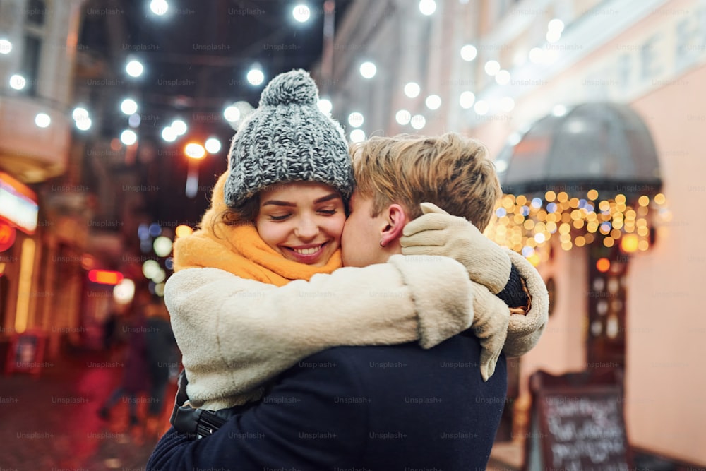 クリスマスで飾られた通りでお互いを抱きしめる暖かい服を着た前向きな若いカップル。