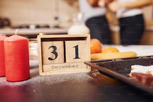 Wooden calendar with 31 december writen on it. Candles and food on the table. People at background.