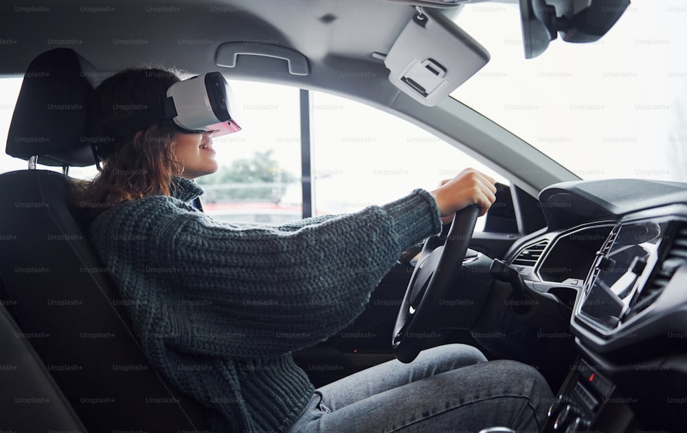 Vue latérale d’une femme dans des lunettes de réalité virtuelle qui est assise à l’intérieur d’une voiture moderne.