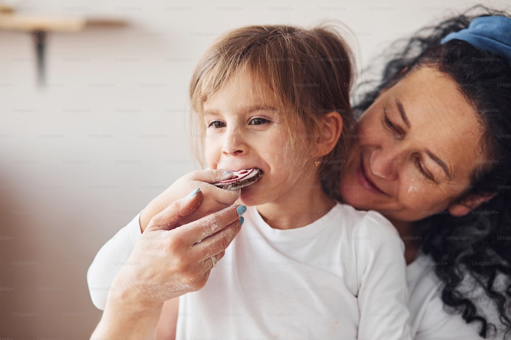 Senior woman with her granddaughter eating fresh cookie on kitchen.