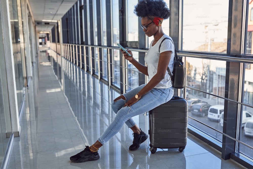 カジュアルな服装の若いアフリカ系アメリカ人の女性通行人は、手荷物を持って空港にいます。電話を使用する。