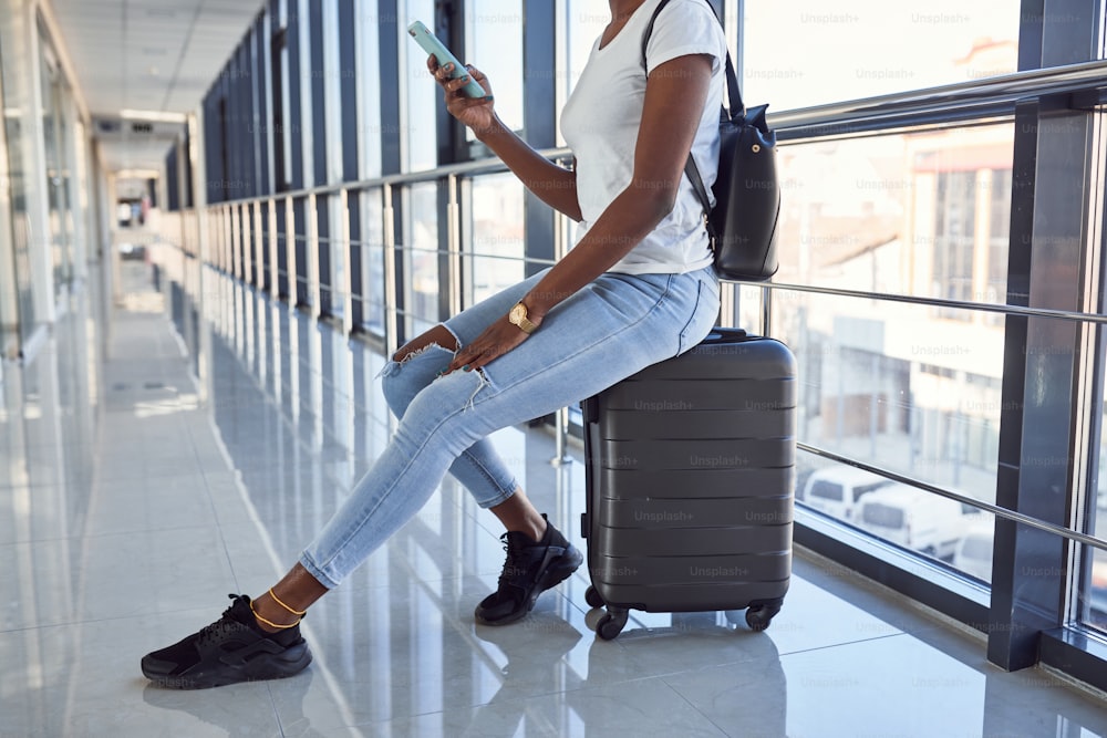 カジュアルな服装の若いアフリカ系アメリカ人の女性通行人は、手荷物を持って空港にいます。電話を使用する。