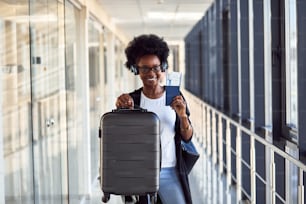 Young african american female passanger in casual clothes and headphones is in airport with baggage.