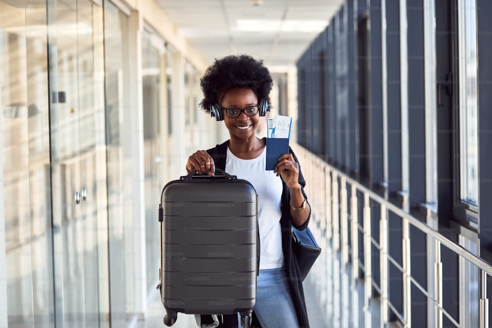 Joven pasajera afroamericana con ropa informal y auriculares está en el aeropuerto con equipaje.
