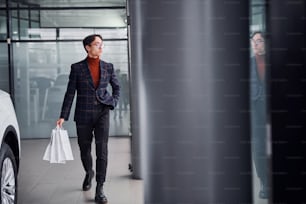 Holding shopping bags. Young business man in luxury suit and formal clothes is indoors near the car.