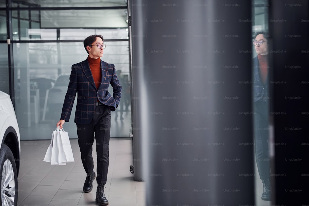 Holding shopping bags. Young business man in luxury suit and formal clothes is indoors near the car.