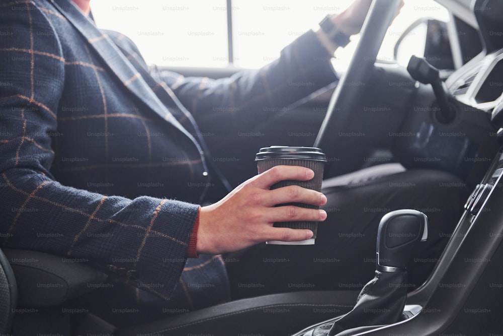 Vista ravvicinata della mano degli uomini con tazza di bevanda. Ragazzo in abito formale seduto all'interno di una nuova auto moderna.